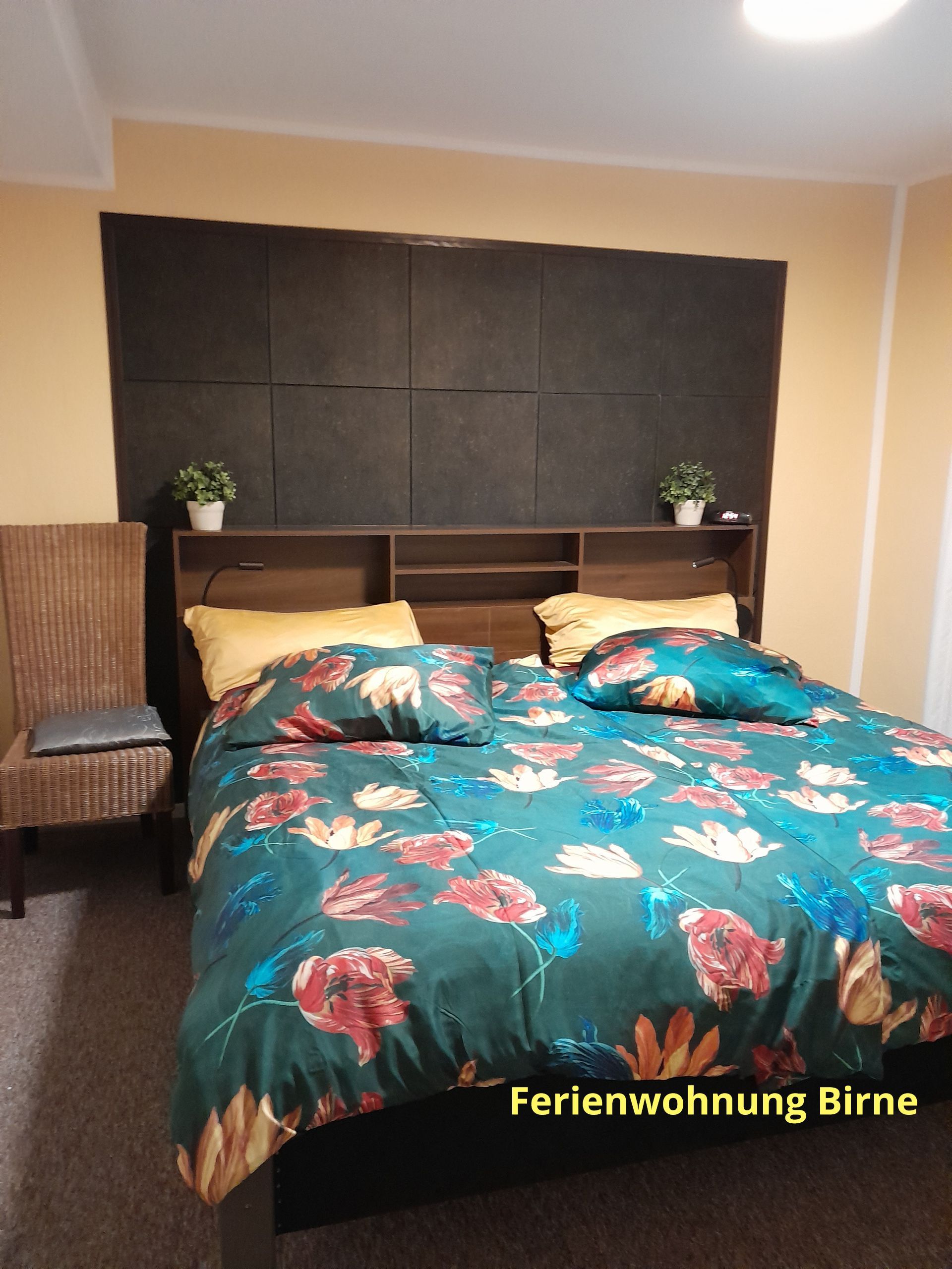 Slaapkamer met bloemenbeddengoed, donker hoofdbord met planken, rieten stoel.