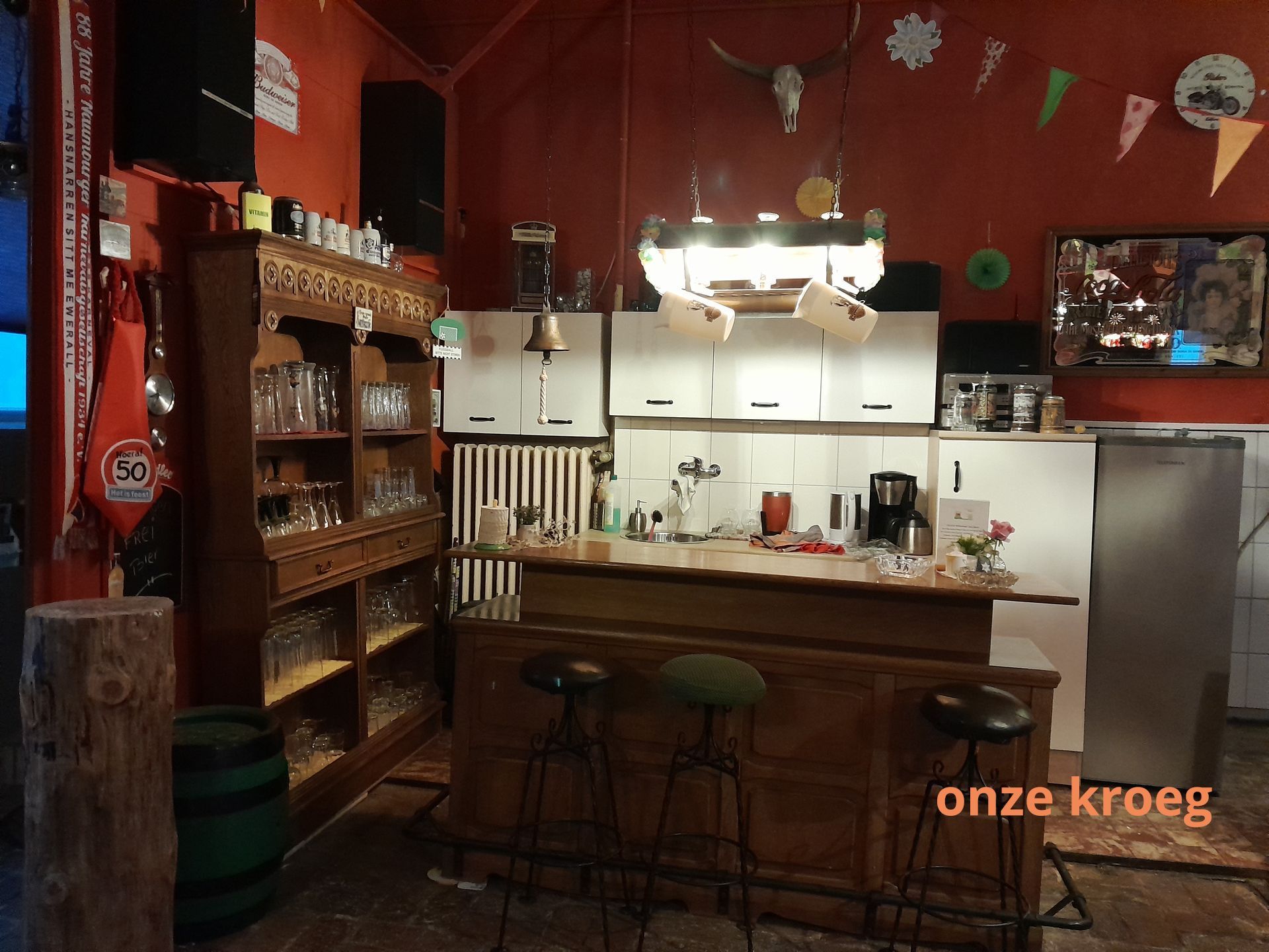 Gezellige bar met houten bar, barkrukken, planken met glazen en rode muren. In de hoek staat 