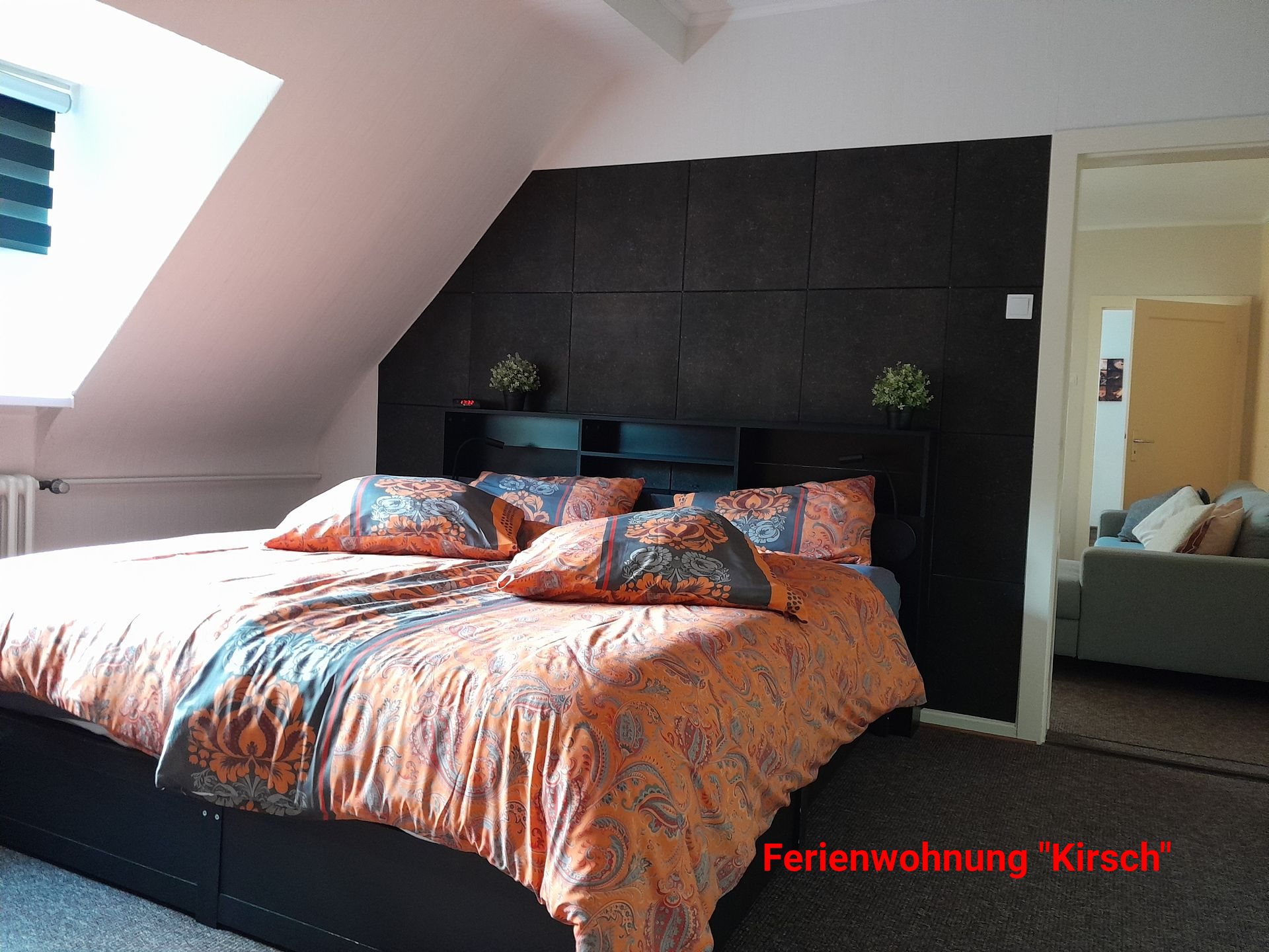 Slaapkamer met bed, donker hoofdeinde, oranje beddengoed en deur naar de woonkamer.