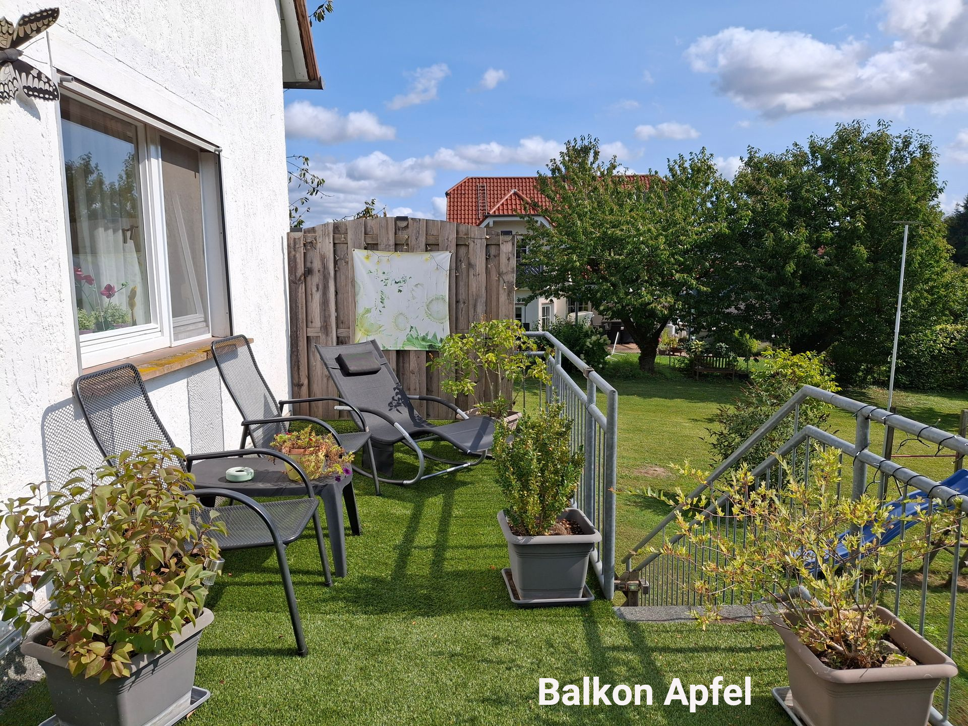 Een balkon met zitjes, potplanten en een tafeltje op een zonnige dag. Een houten schutting en bomen zijn zichtbaar.