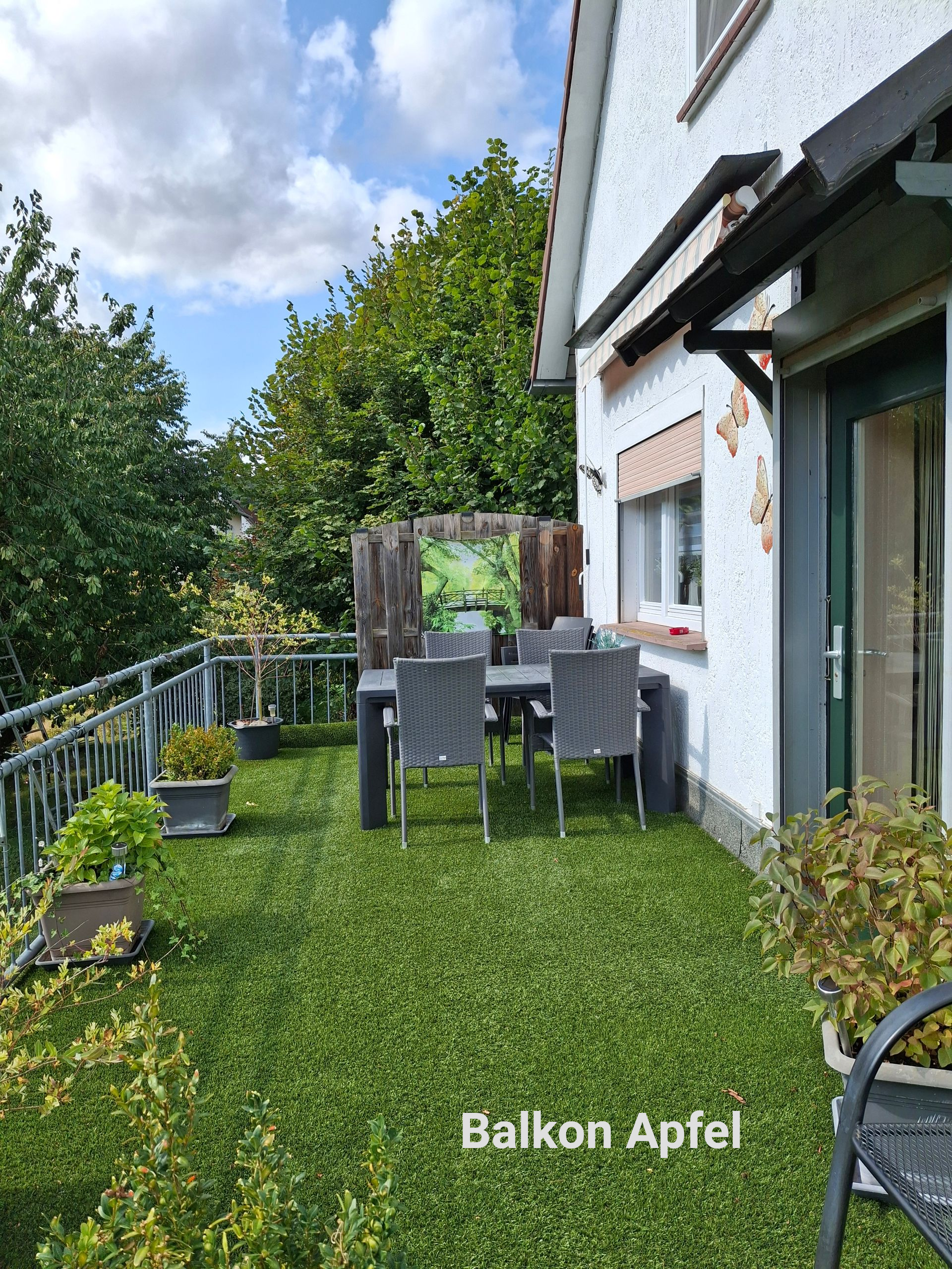 Een balkon met kunstgras, een tafel en stoelen. De muur van een wit huis is zichtbaar. Groene bomen en planten omringen het huis.