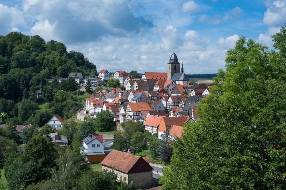 Een stadje verscholen tussen bomen, met rode daken, een kerktoren en een blauwe hemel met pluizige wolken.