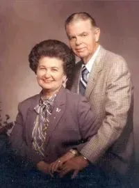 A studio portrait of a couple. The man stands behind the seated woman, both smiling against a soft, neutral background.