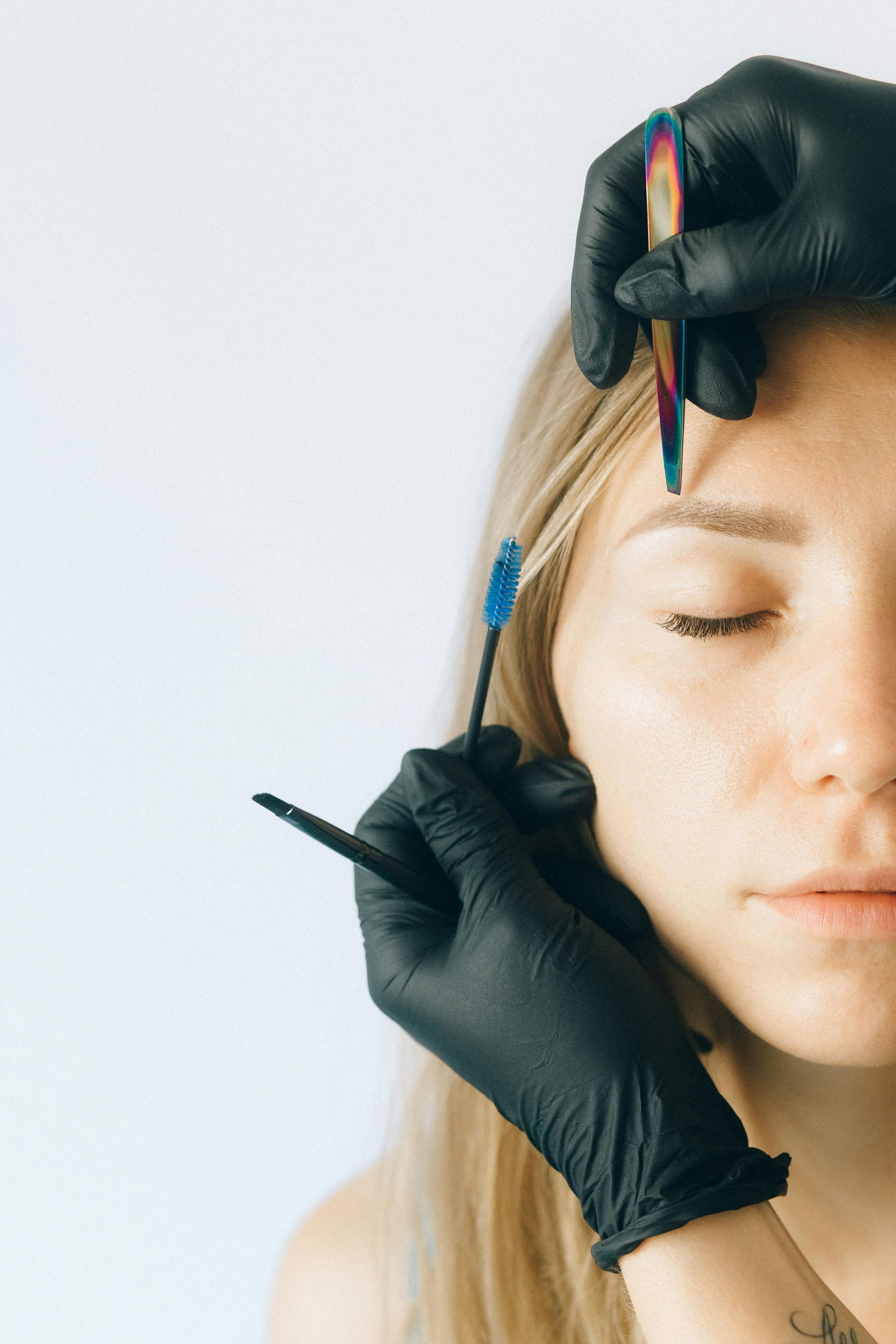 Woman's eyebrow being shaped by gloved hands with a brush and pencil.