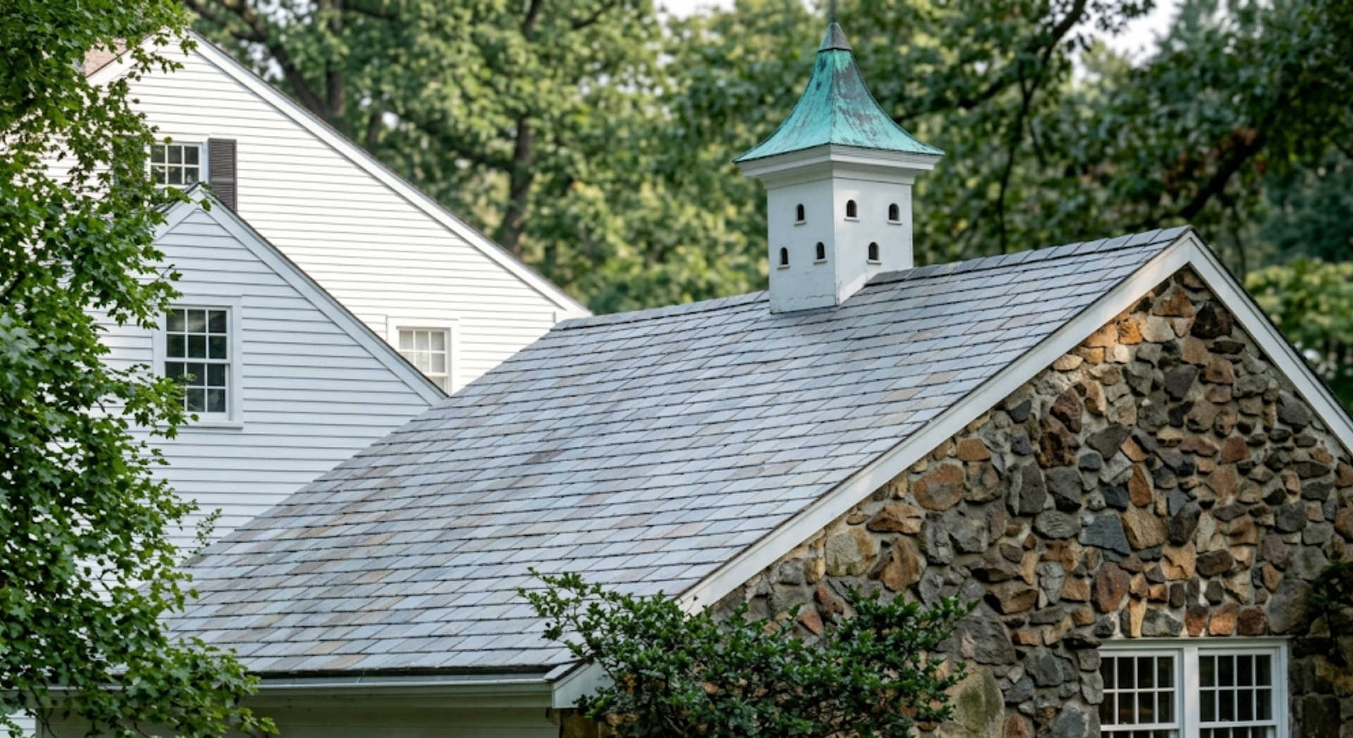 Stone house roof with white siding and a small cupola, surrounded by trees