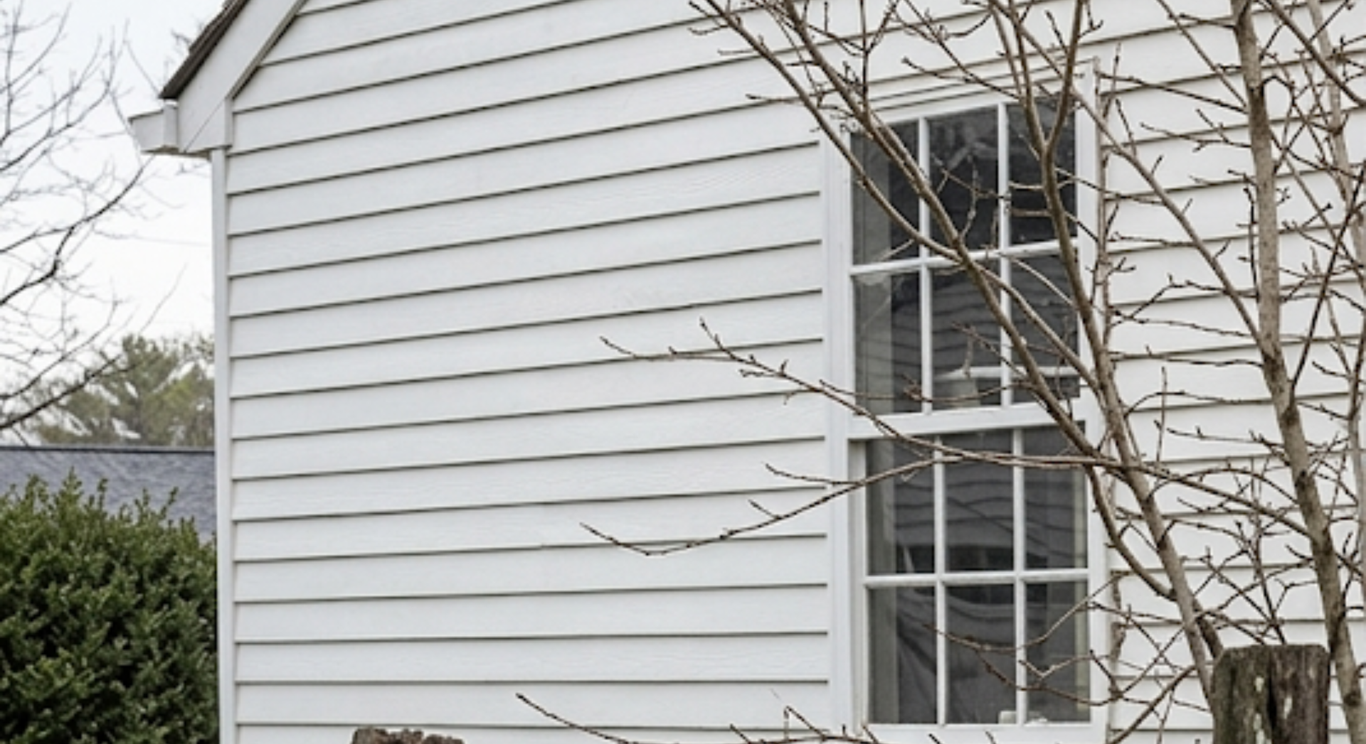 White house siding with a tall window partly obscured by bare tree branches and a hedge.
