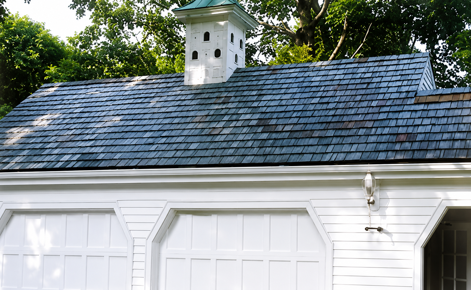 White building with a gray shingled roof and small cupola, surrounded by green trees