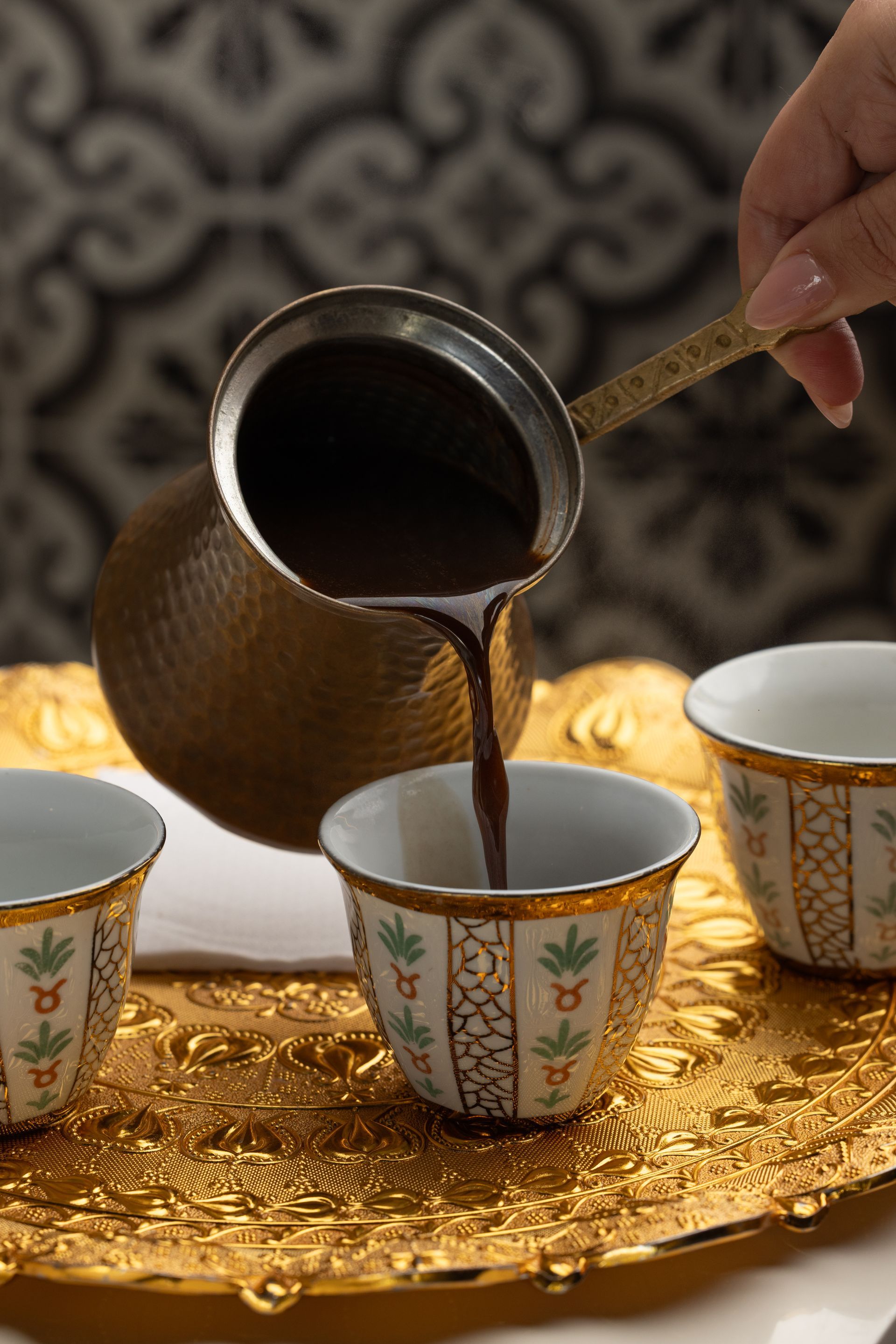 Coffee being poured from a copper pot into a small cup, on a gold tray.