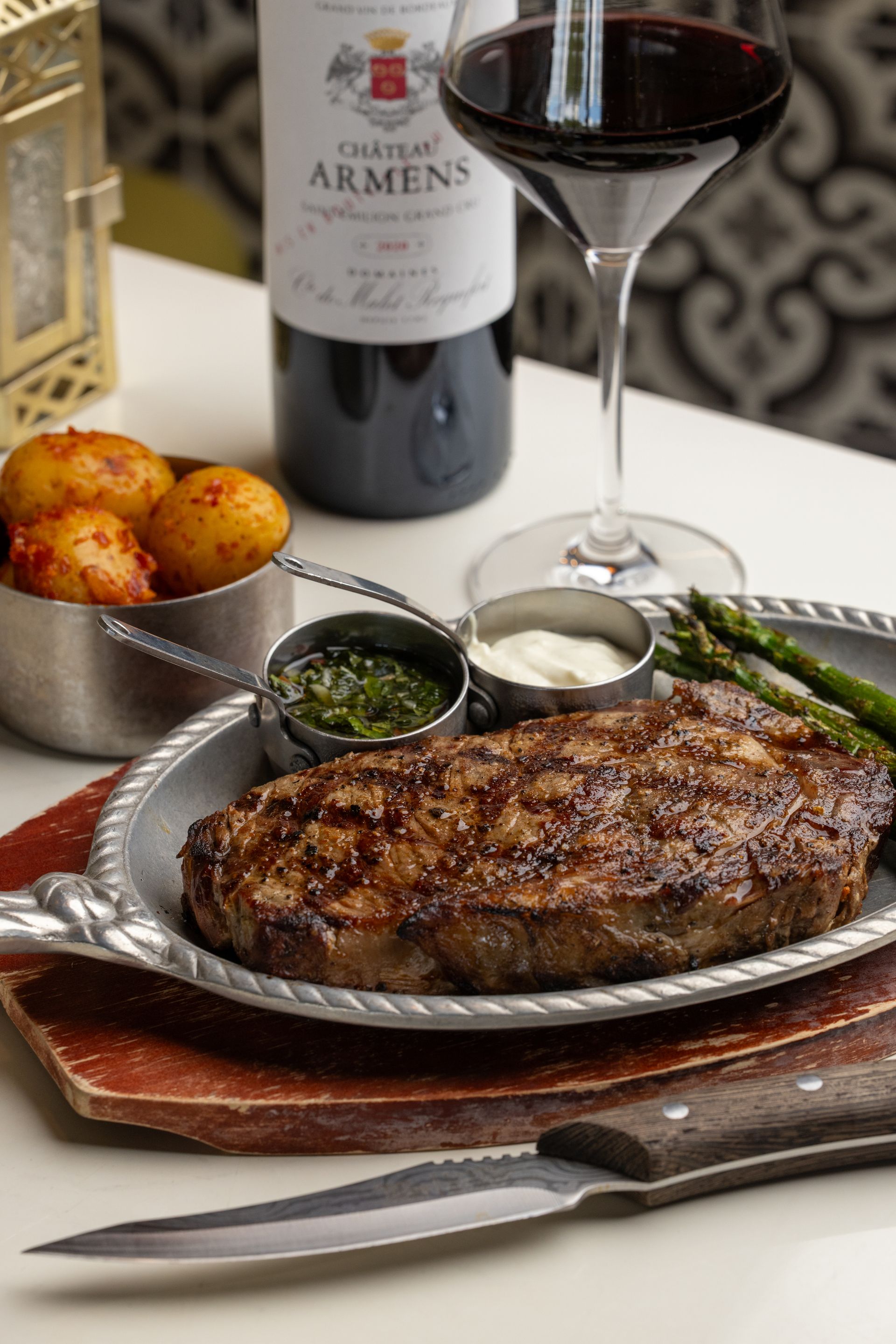 Grilled steak with dipping sauces, asparagus, roasted potatoes, red wine, and a wine glass.