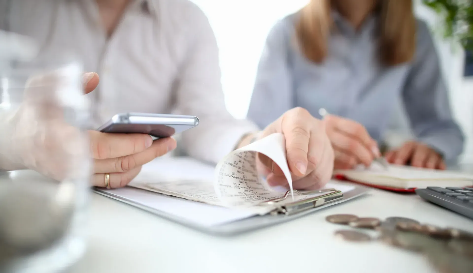 Hands counting cash on a notebook beside a calculator, with a phone and coins on a table.
