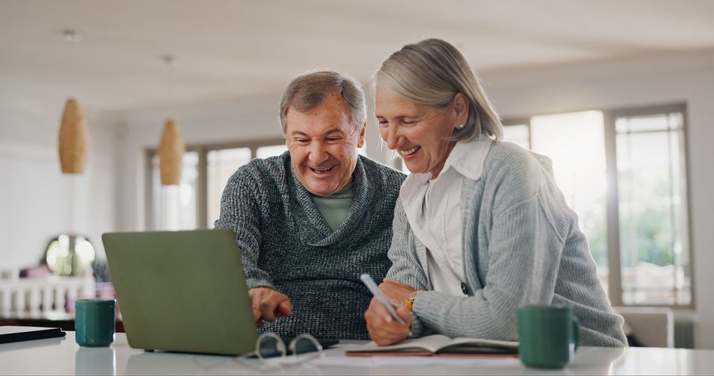 Two people smiling at a laptop in a bright kitchen, with mugs on the table