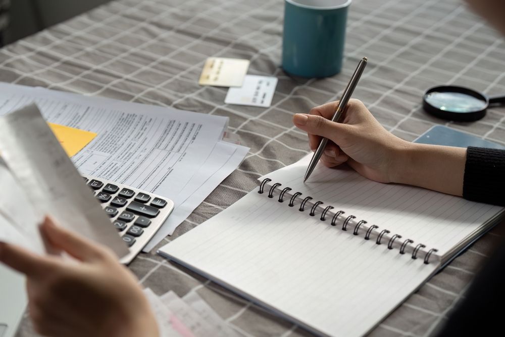 Hands writing in a spiral notebook beside papers, a calculator, and a cup on a desk