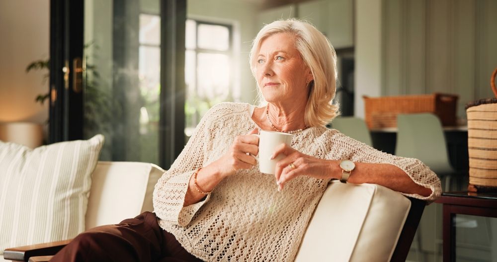 Older person sitting on a sofa, holding a mug and gazing out a sunlit window