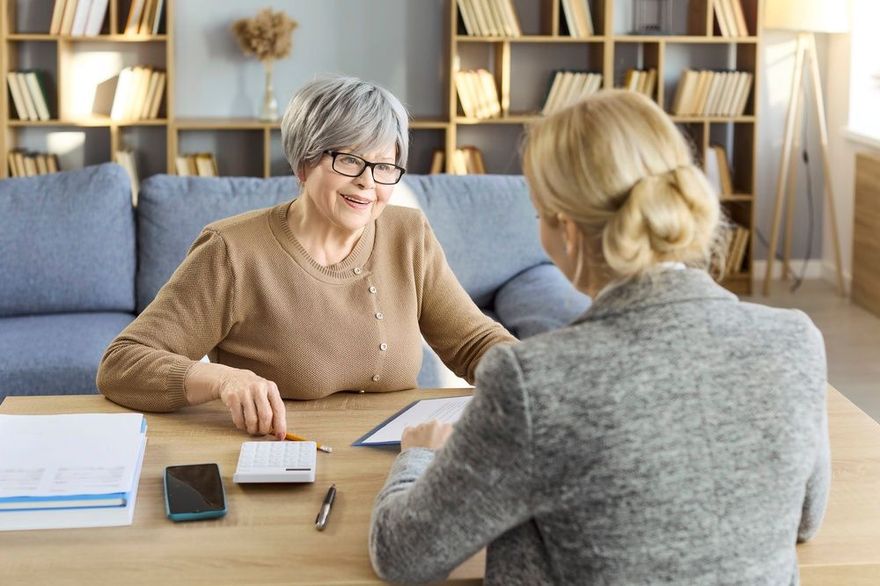 Two people talking at a table in a cozy office, with papers, a calculator, and bookshelves behind them