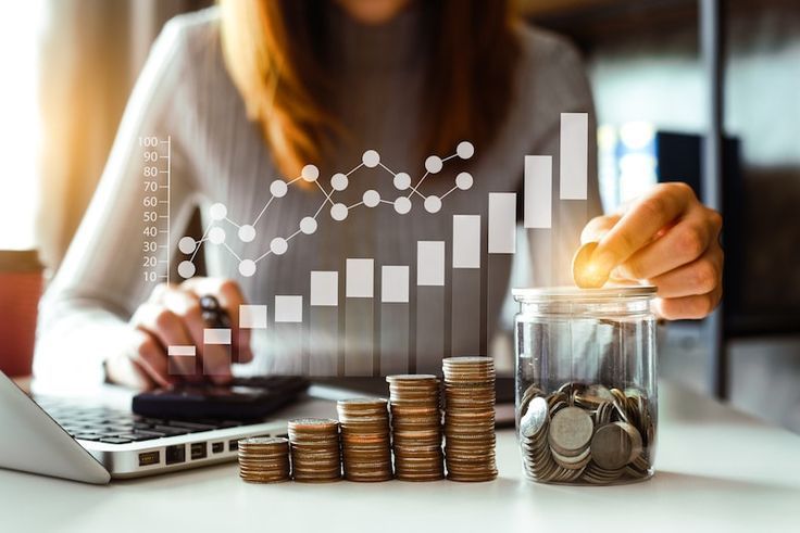 Hand placing coin on laptop beside stacked coins and financial chart overlay