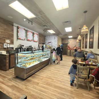Interior of a cafe with pastry display, customers, and tables. Wooden floors, decorative wall art, and warm lighting.