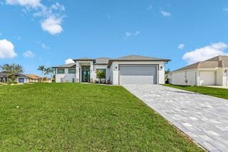 White house with a gray garage door, green lawn, blue sky, and a paved driveway.