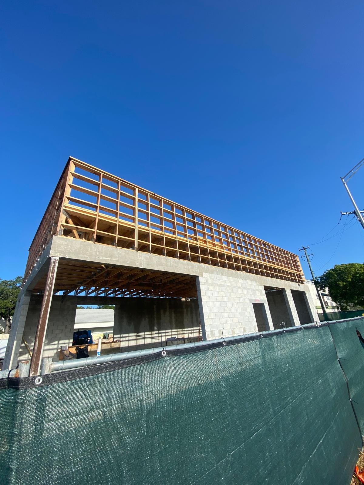 Building under construction with wooden frame, concrete walls, and green fence, against a blue sky.