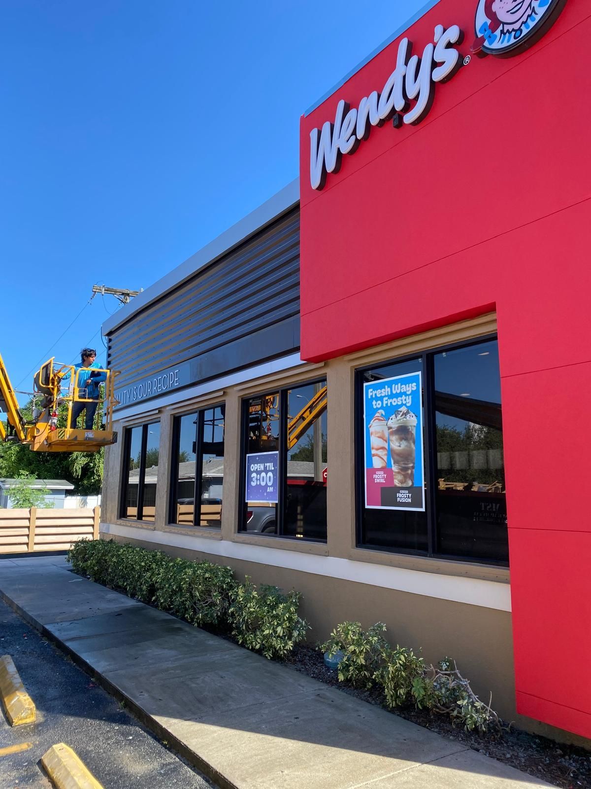 Wendy's restaurant exterior with worker in lift. Red building, windows, signage, and shrubbery. Blue sky.