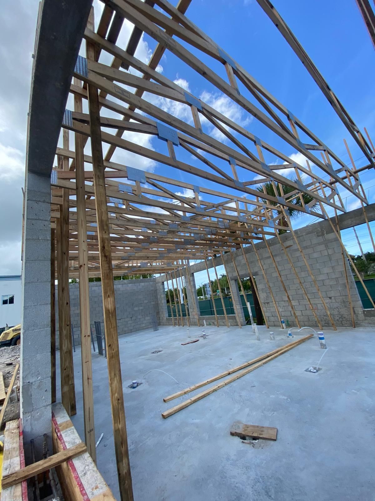 Construction site with exposed wooden roof trusses against a partly cloudy sky. Concrete walls and floor.