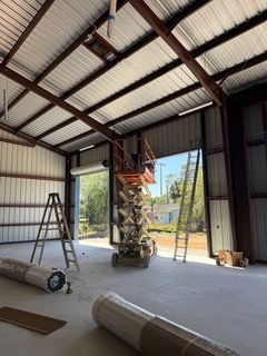 Interior of a warehouse under construction with a lift, ladders, and rolled materials on a concrete floor.