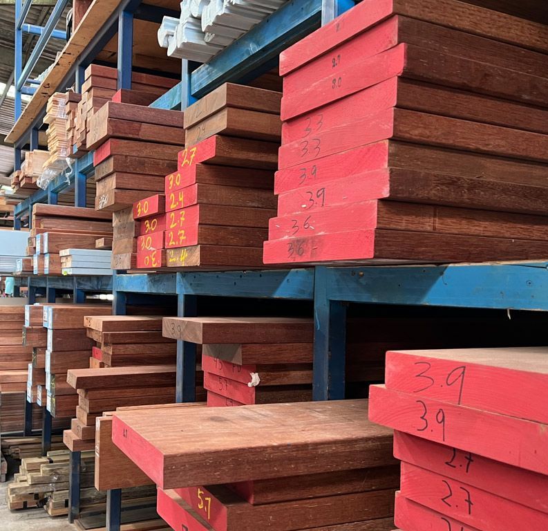 Stacks of cut lumber on blue metal racks in a warehouse, with red-painted ends and handwritten labels. — B & B Timbers Ballina In West Ballina, NSW