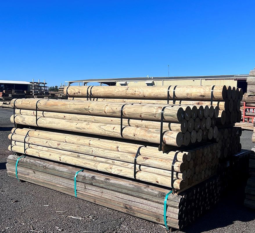 Stacked wooden utility poles on a gravel lot under a clear blue sky. — B & B Timbers Ballina In West Ballina, NSW