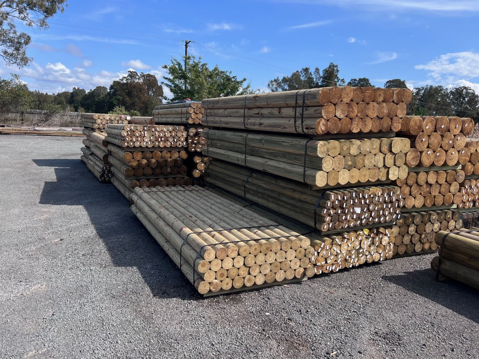 Stacks of lumber logs and timber planks on a gravel lot under a blue sky. — B & B Timbers Ballina In West Ballina, NSW