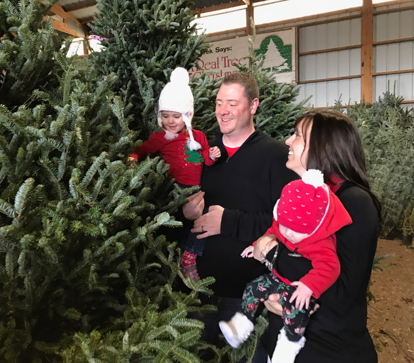 A family smiles while picking out a Christmas tree in a nursery, with two young children in winter hats and red sweaters.