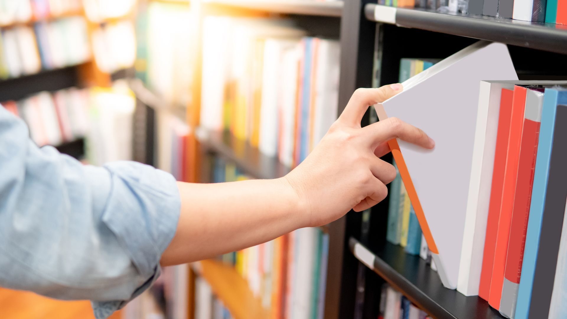 Person's hand reaching for a book on a library shelf filled with colorful books.