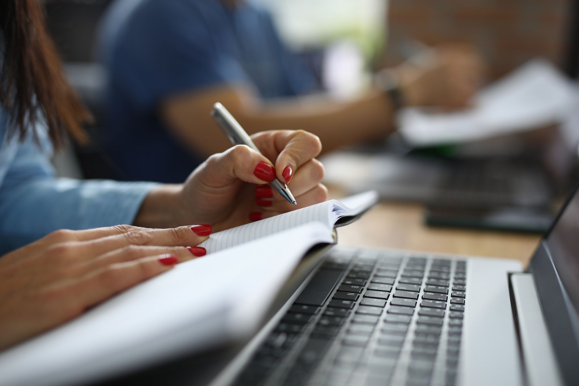 Woman with red nails writes in a notebook, next to a laptop. Another person is in the blurred background.