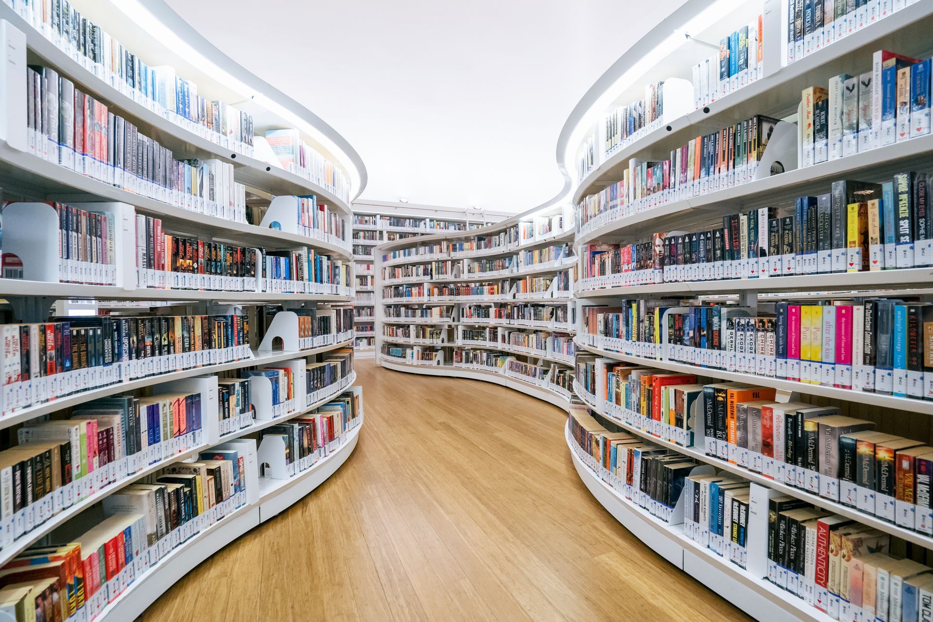 Curved bookshelves filled with books in a library, light wood floor, white ceiling.