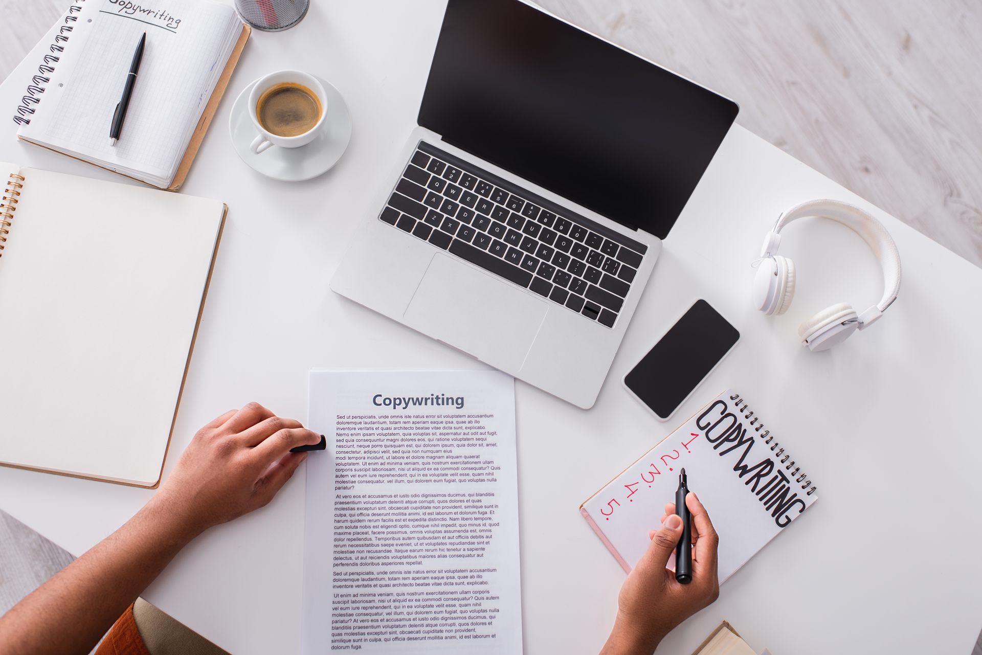 Hands writing copy on a notepad next to a laptop, documents, coffee, and headphones on a white desk.