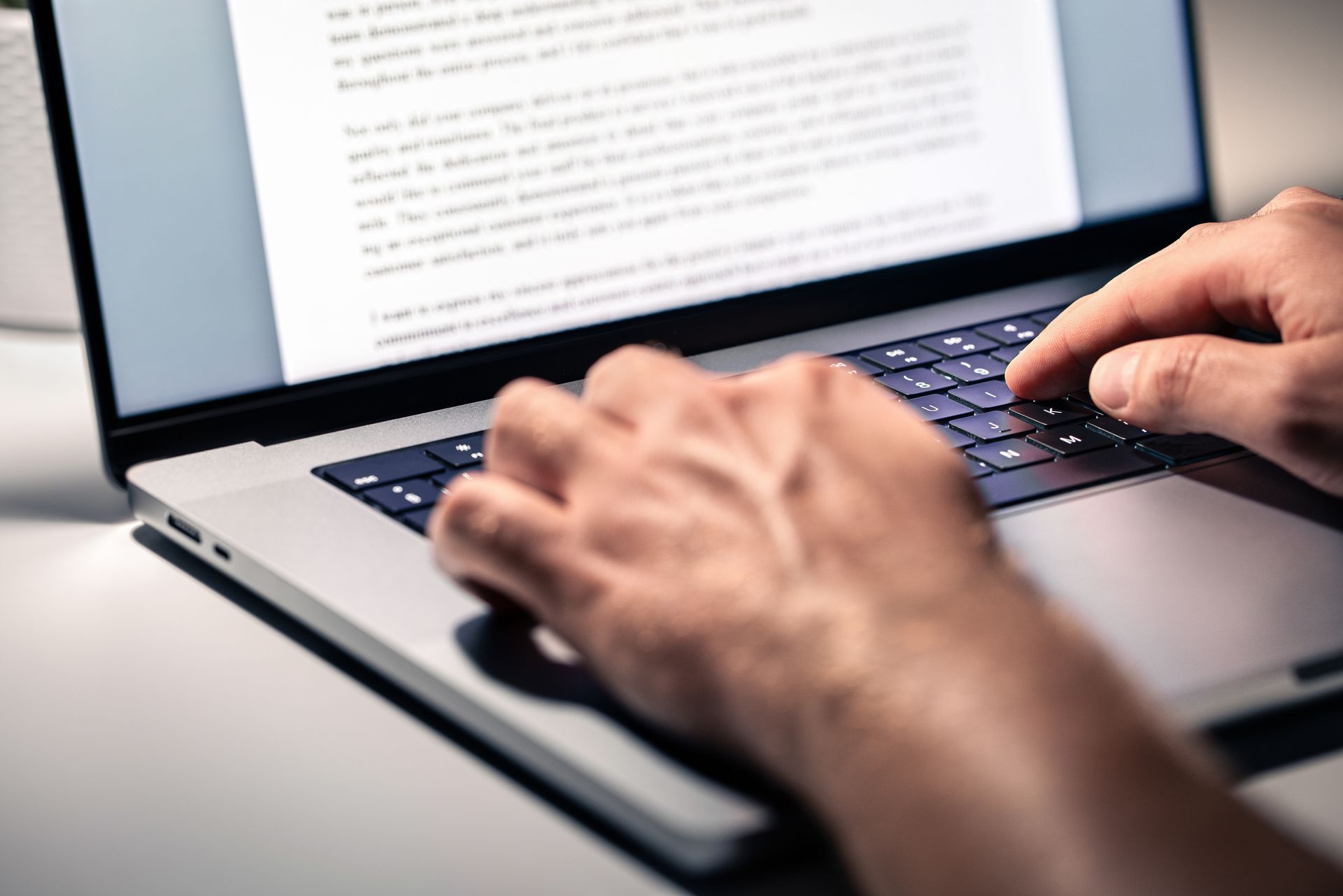 Hands typing on a laptop keyboard; screen displays document on a white desk.