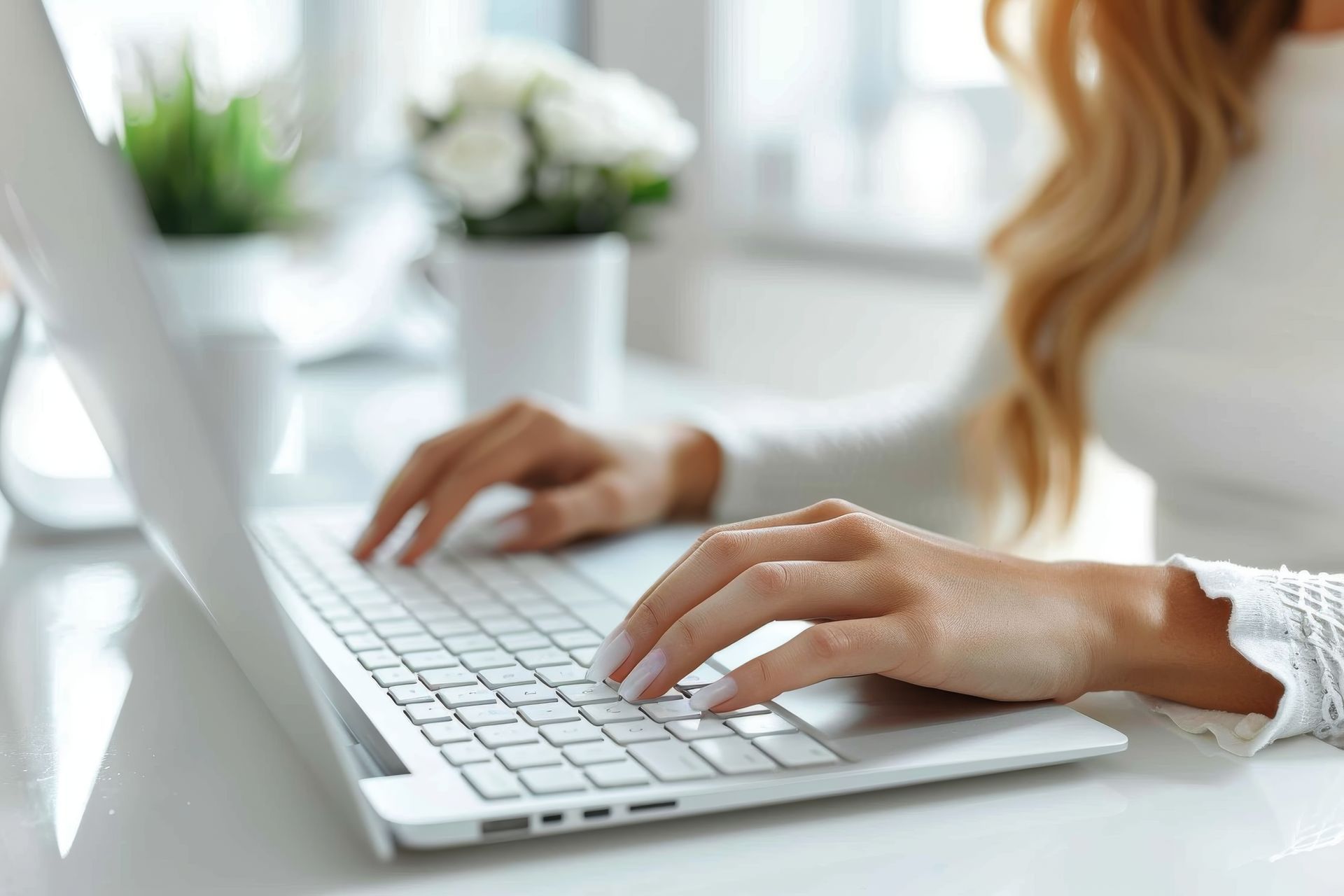 Woman typing on a white laptop, blurred background with flowers and plants.