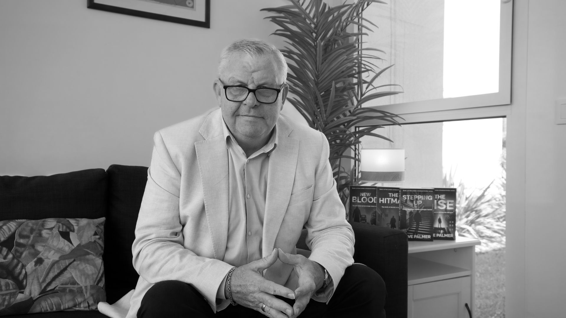 Man wearing glasses and a light-colored blazer sitting on a couch, indoors.