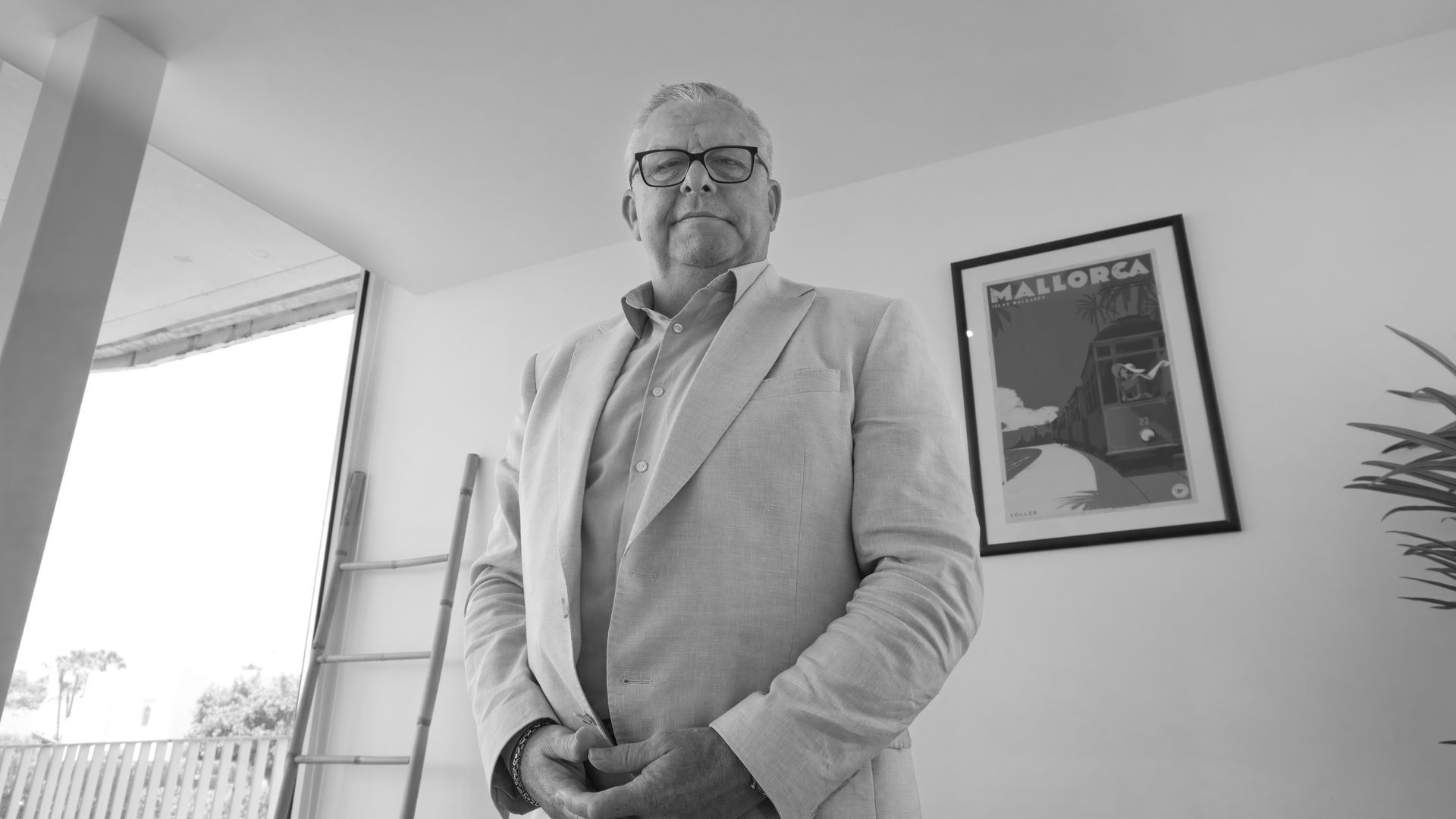 Man in a light suit standing by a window in a bright office, with framed art on the wall.
