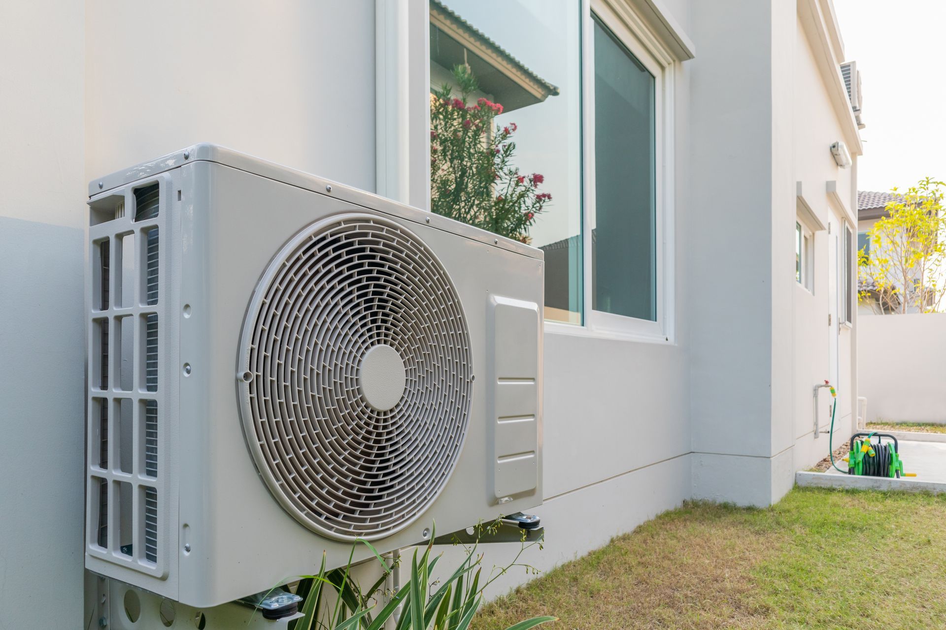 Outdoor air conditioner unit beside a white house with windows and a small lawn