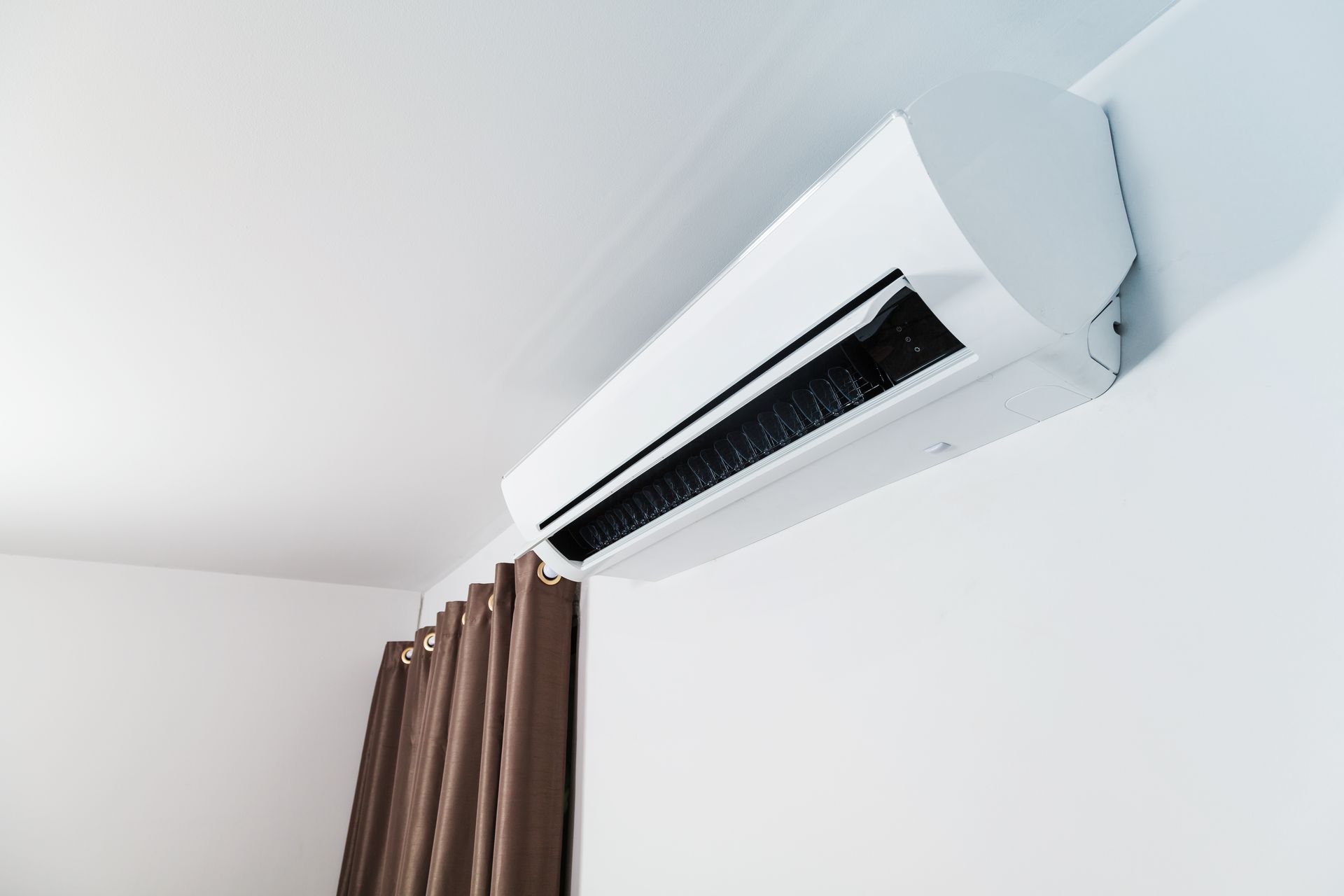 Wall-mounted white air conditioner above brown curtains in a white room