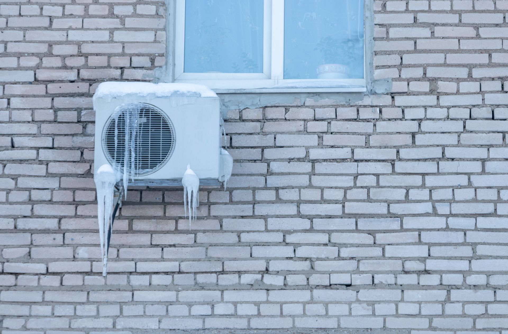 Snow-covered air conditioner mounted on a brick wall below a window.