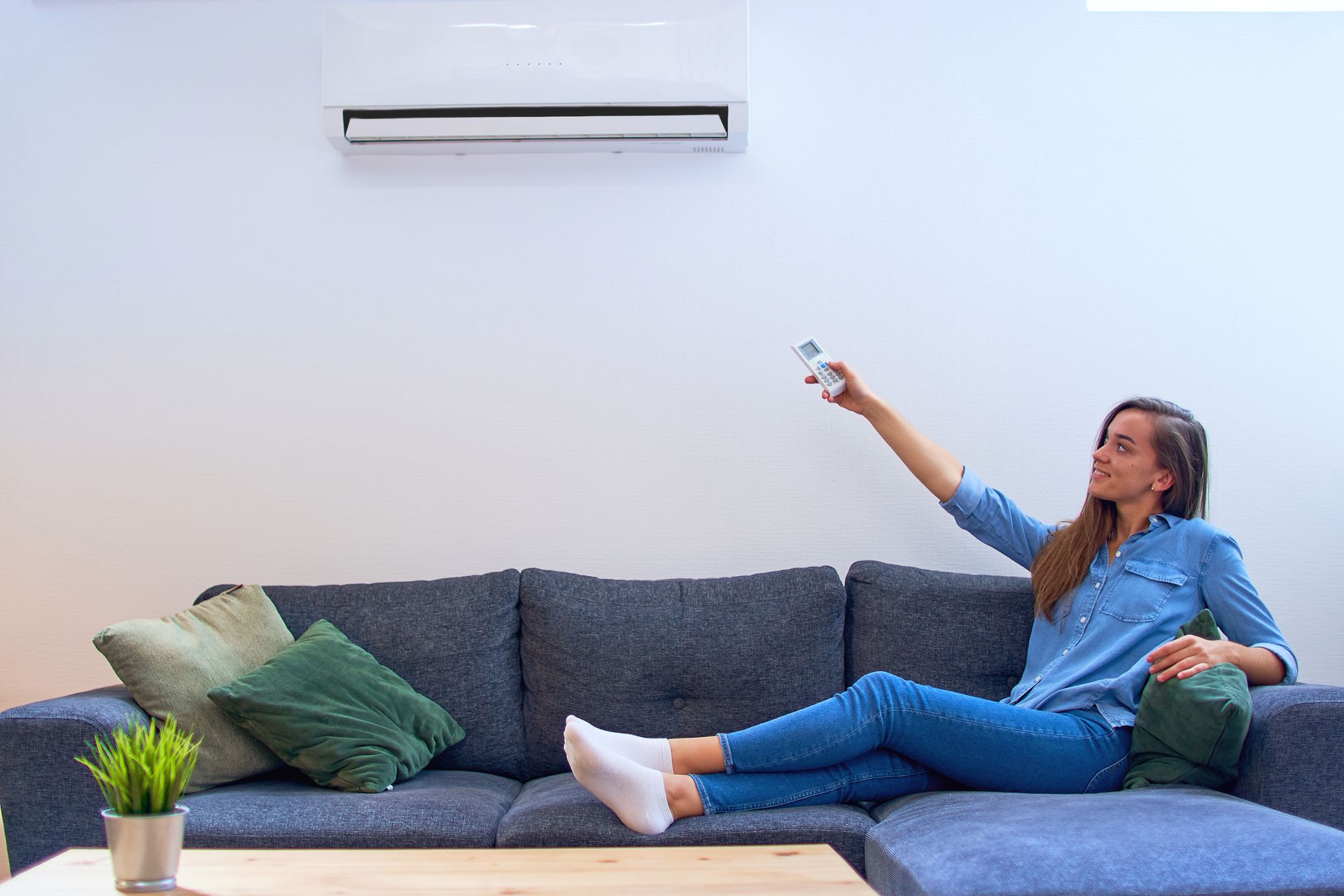 Woman lounging on a blue sofa, pointing a remote at a wall-mounted air conditioner in a bright living room