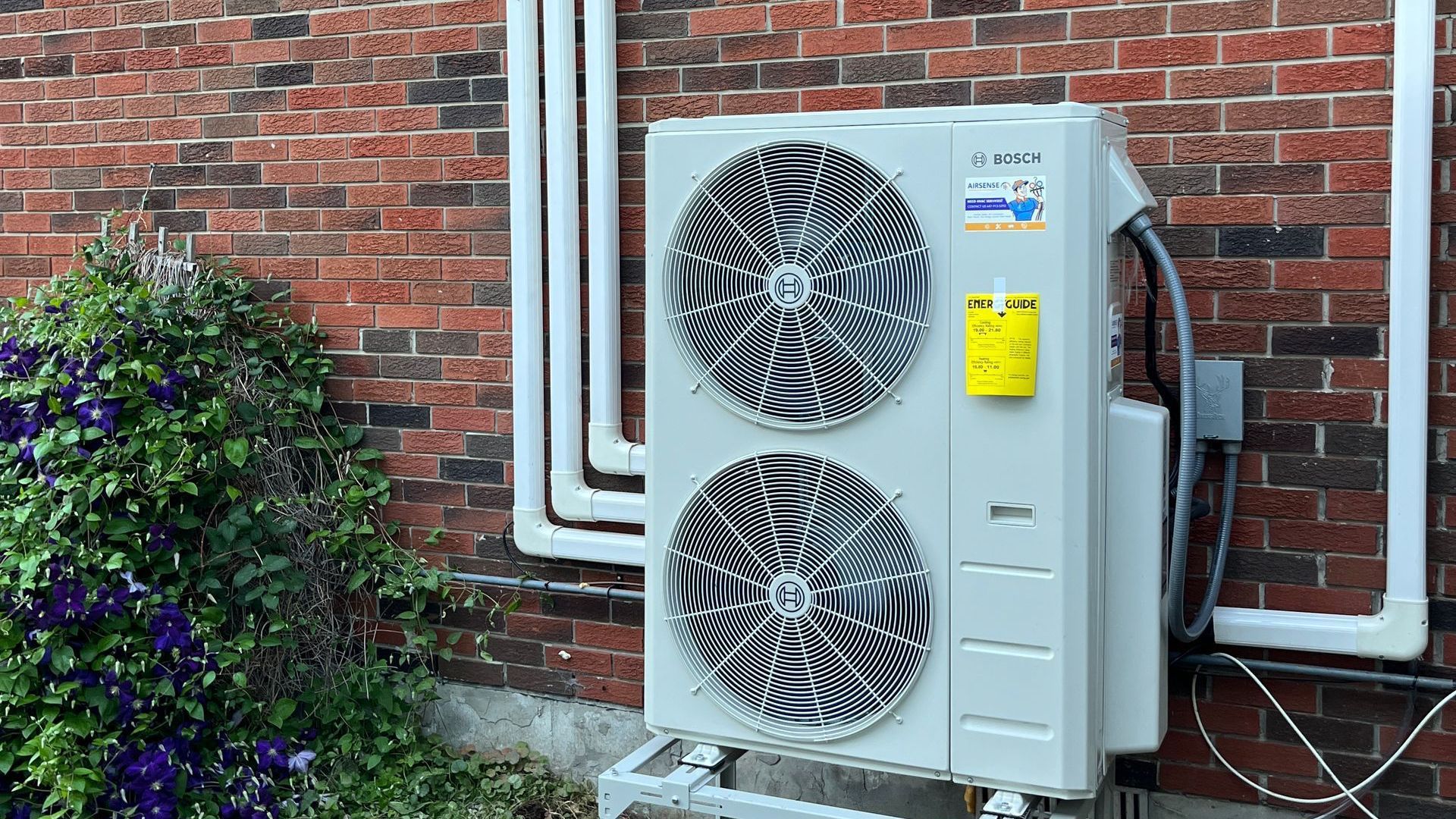A white air conditioner is sitting outside of a brick building next to a hose.