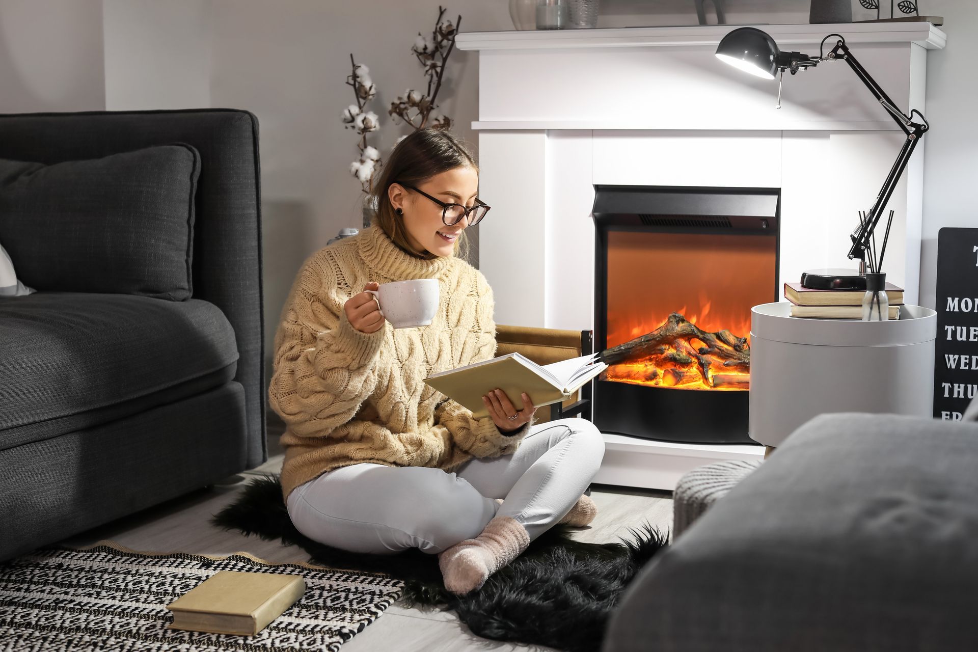 A woman is sitting on the floor reading a book in front of a fireplace.