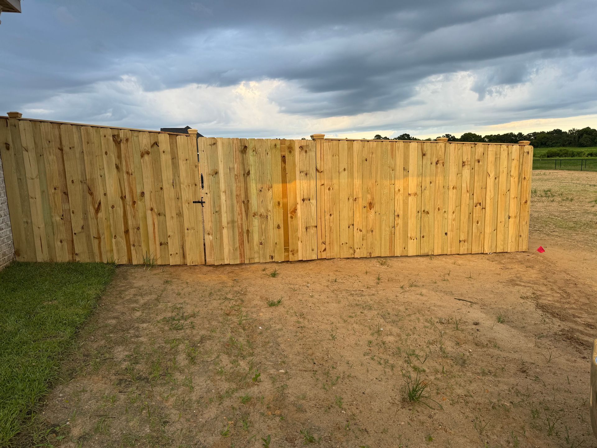 Wooden privacy fence against a cloudy sky, bordering a dirt yard and grass.