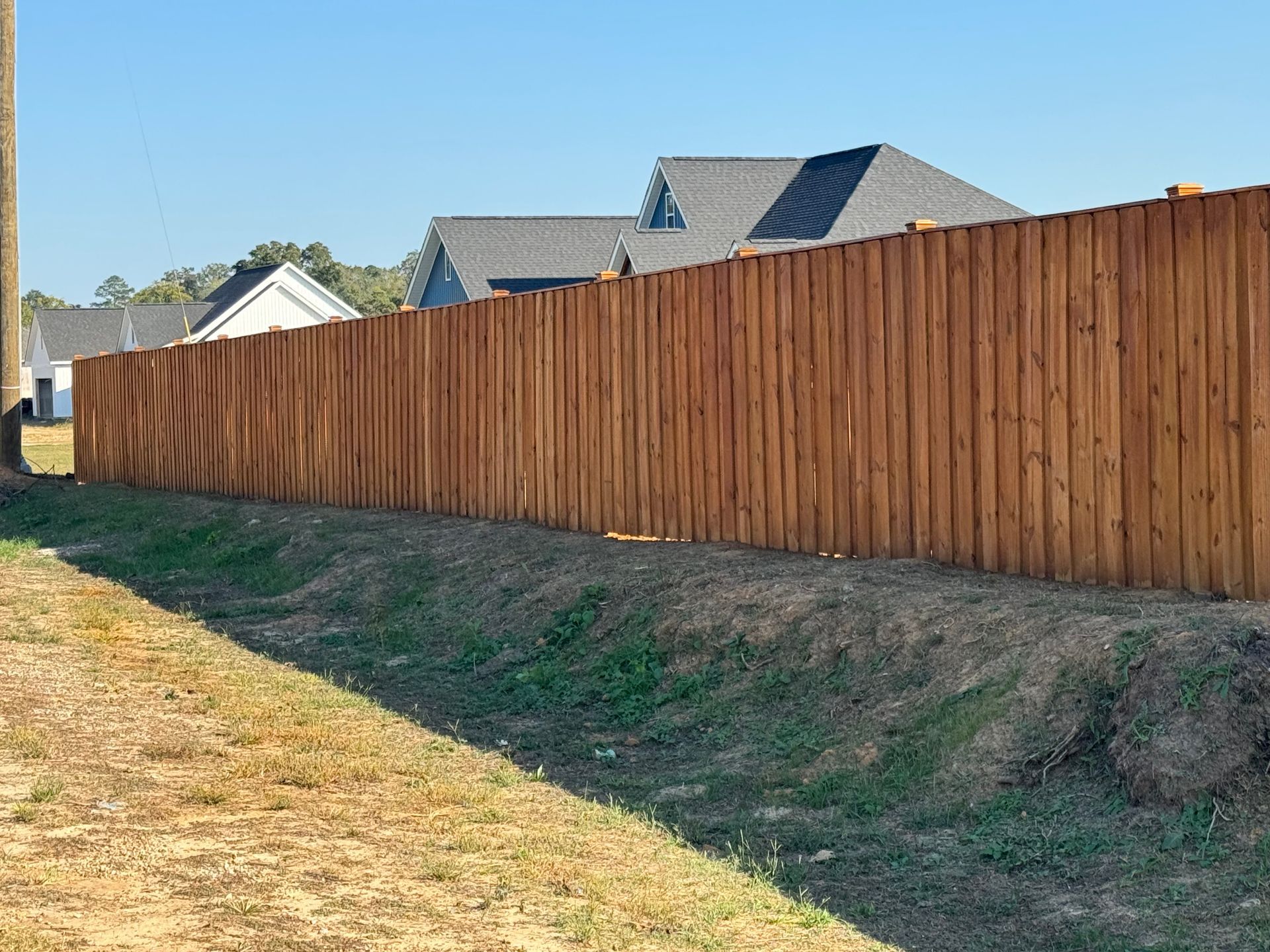 Wooden fence along a grassy verge on a sunny day. Houses visible in the background.