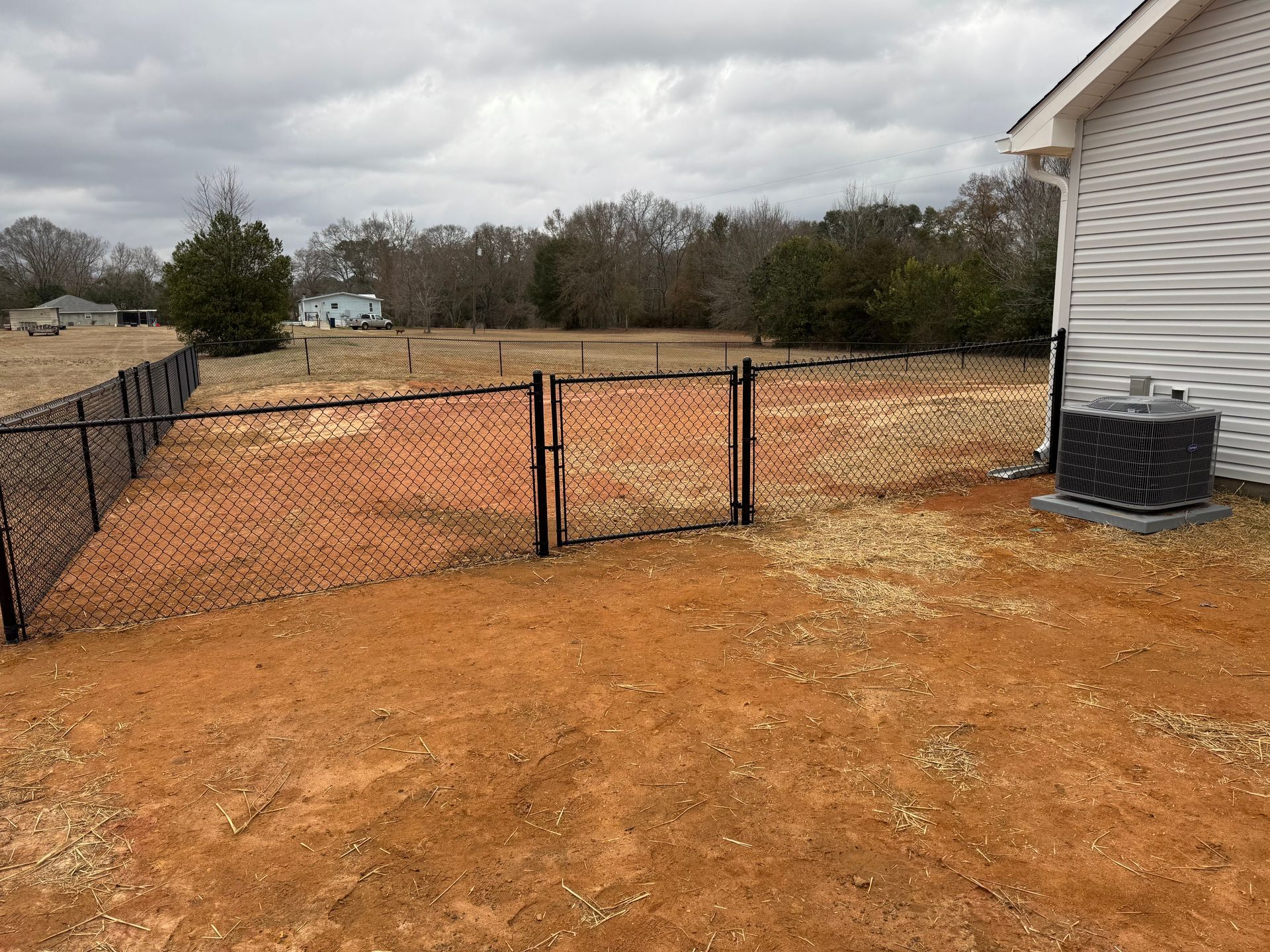 Black chain-link fence and gate in a yard with red soil, next to a white house with a visible AC unit.