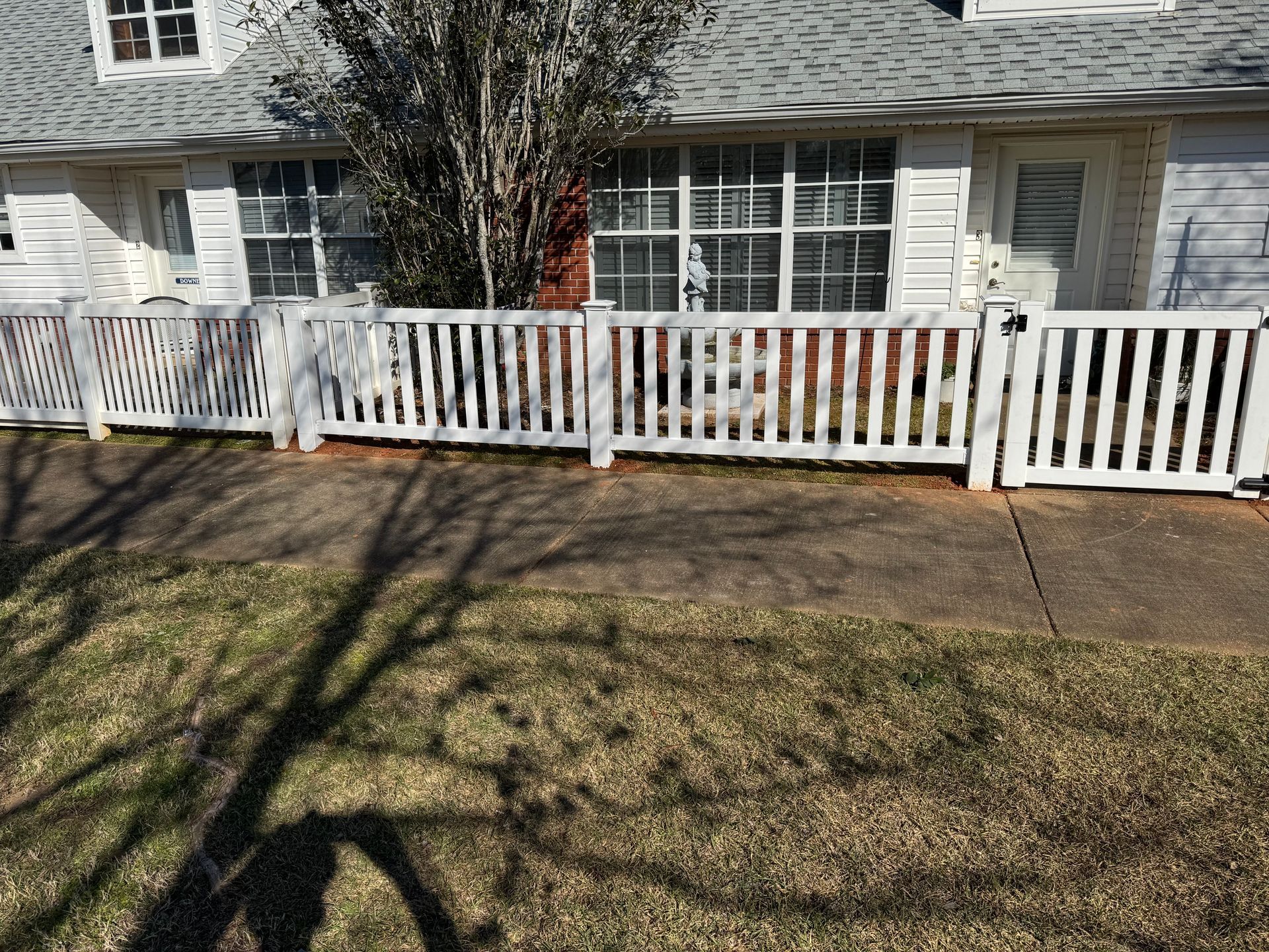 White picket fence in front of a white house with multiple windows and a gray roof; a brown tree.