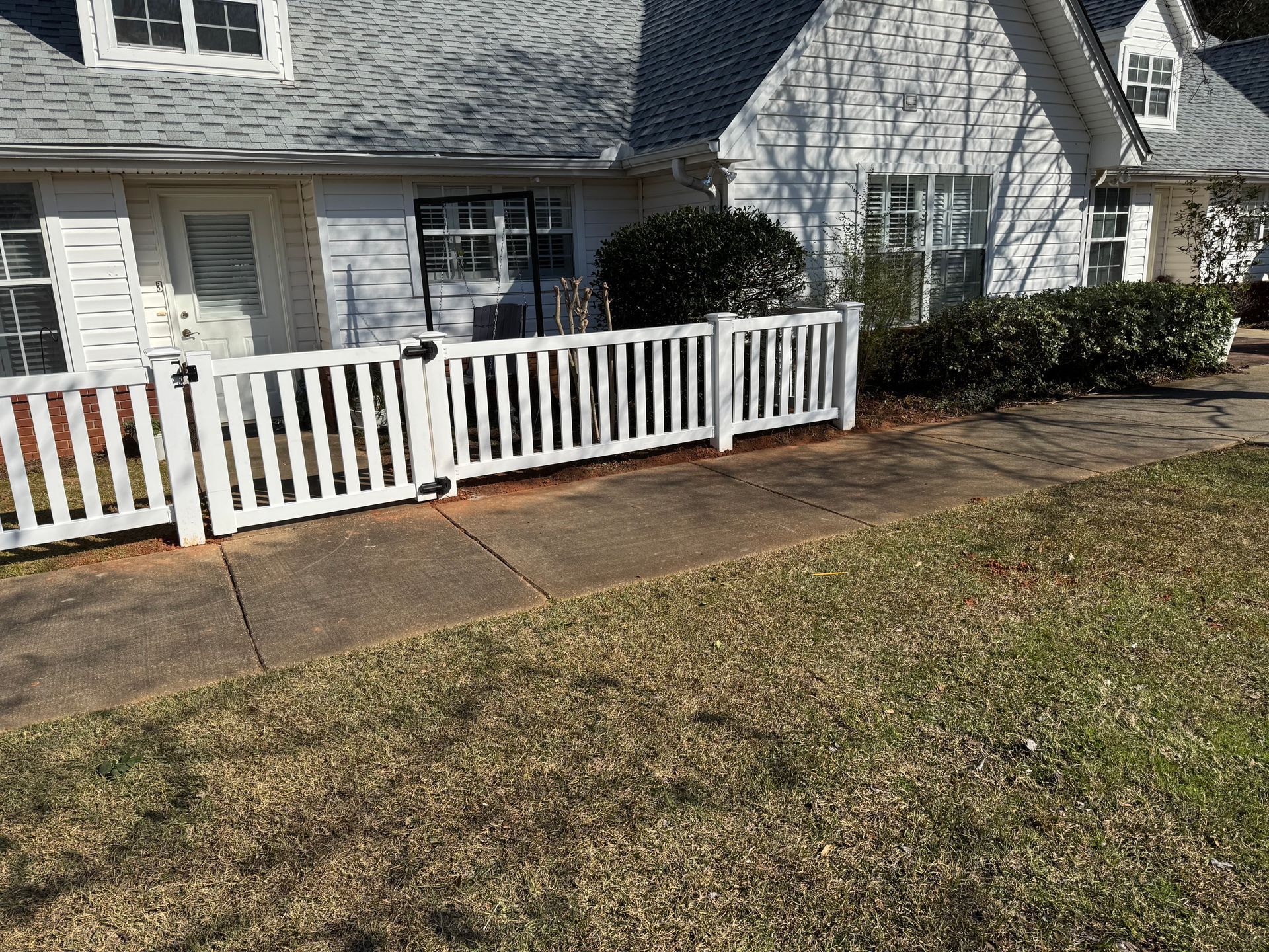 White picket fence in front of a white house with a gray roof and shrubs. Concrete sidewalk and grass in view.