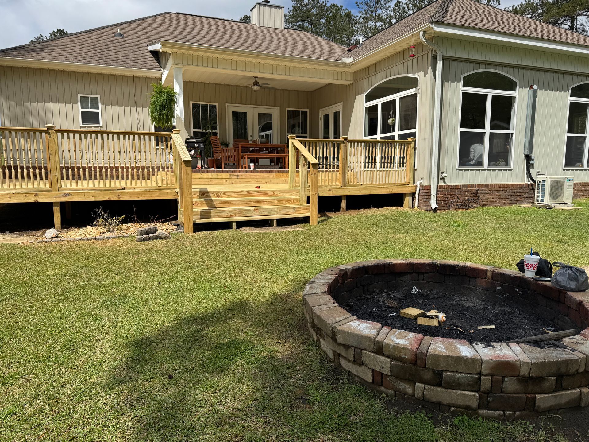Wooden deck and brick fire pit in a backyard with green grass, next to a light-colored house.