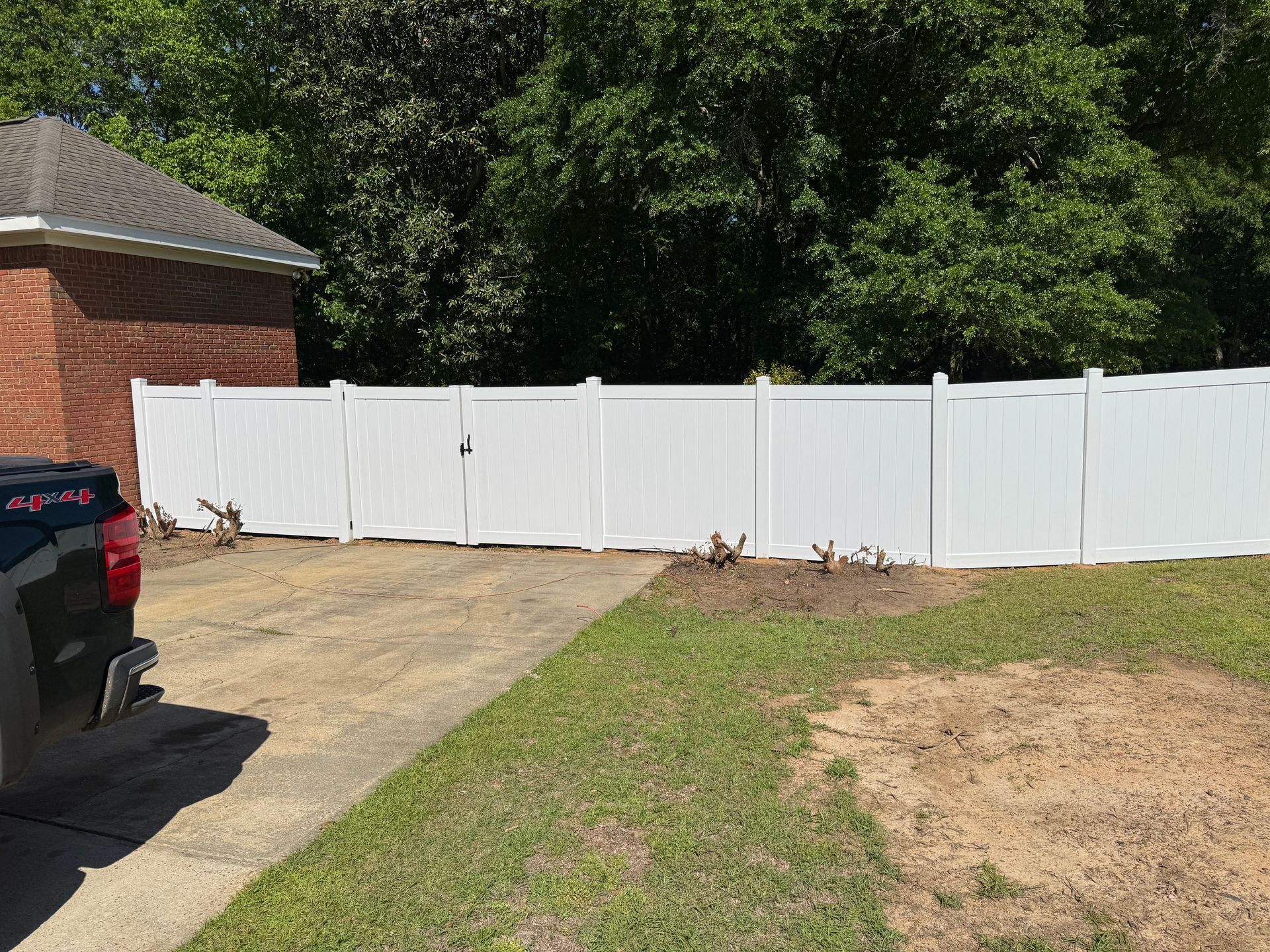 White vinyl fence in a yard next to a driveway and a brick house. Green grass and trees are in the background.